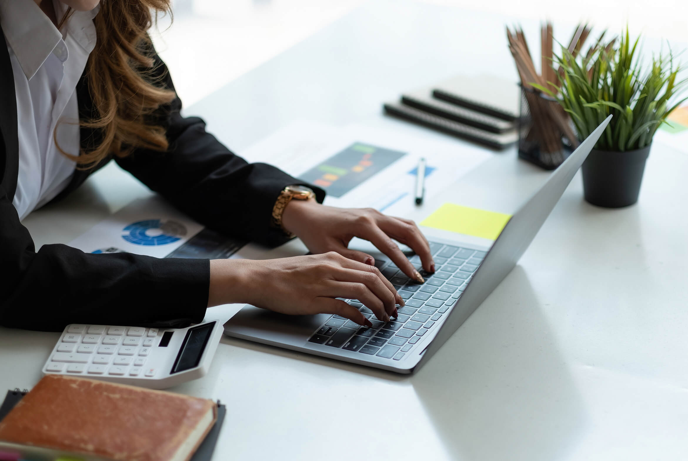 A woman with long hair in a black blazer types on a laptop at a desk, with a calculator, papers, a notebook, pens, and a potted plant nearby.