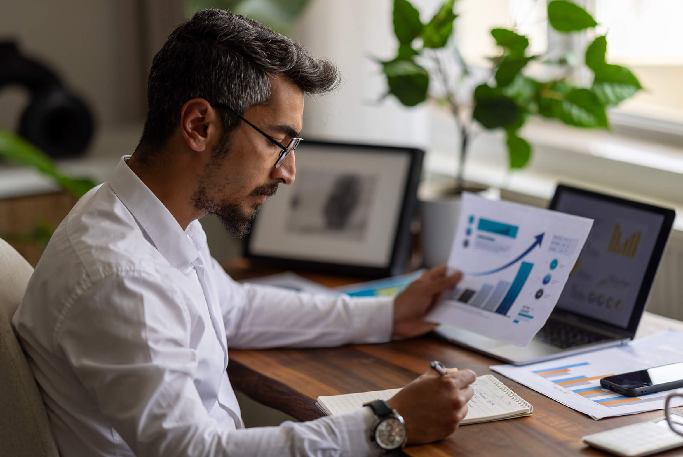 A man with short, dark hair wearing glasses and a white shirt is working at a wooden desk. He is holding a paper with charts and graphs, while writing in a notebook with a pen. A laptop displaying similar charts, a smartphone, and a potted plant are on the desk. The background includes a framed picture and a window with natural light.