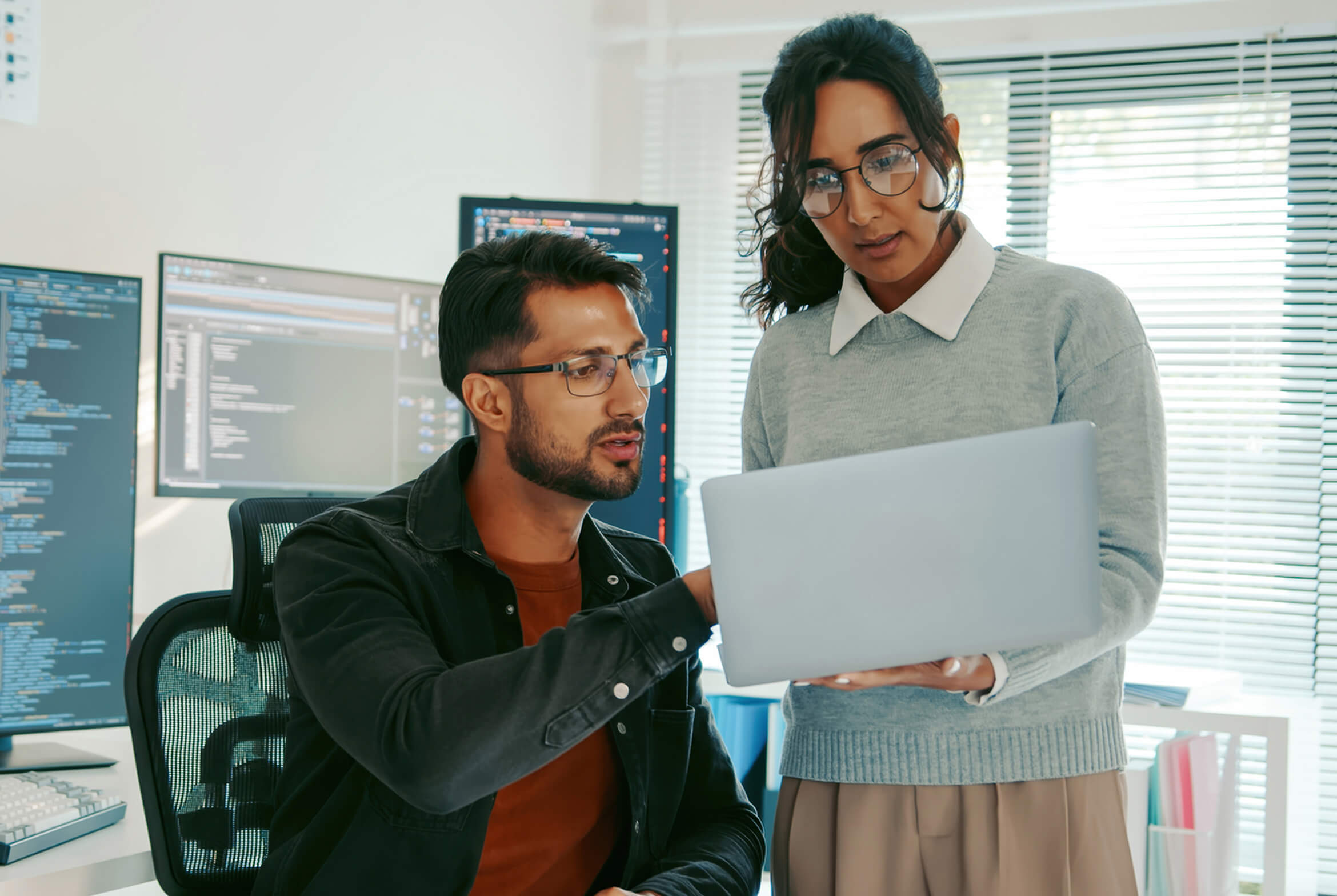 A man and a woman collaborating in an office, with the woman holding a laptop and the man seated at a desk with multiple monitors displaying code.