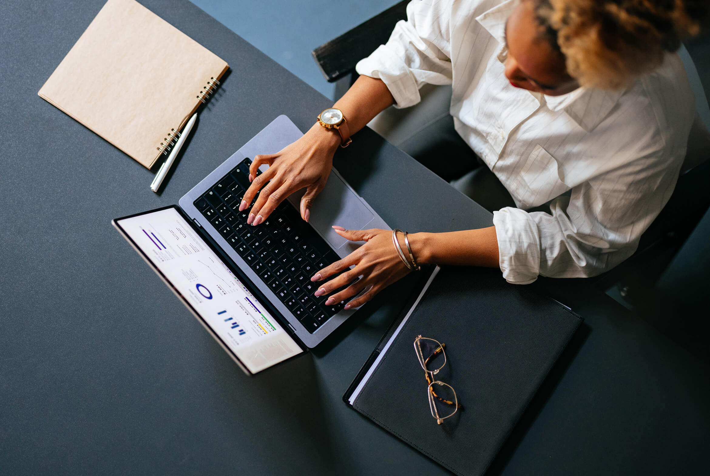 A person with curly hair is working on a laptop at a desk. The laptop screen displays charts and data. On the desk, there are a notebook with a pen, a pair of glasses resting on a closed folder, and the person is wearing a white shirt, a watch, and bracelets.