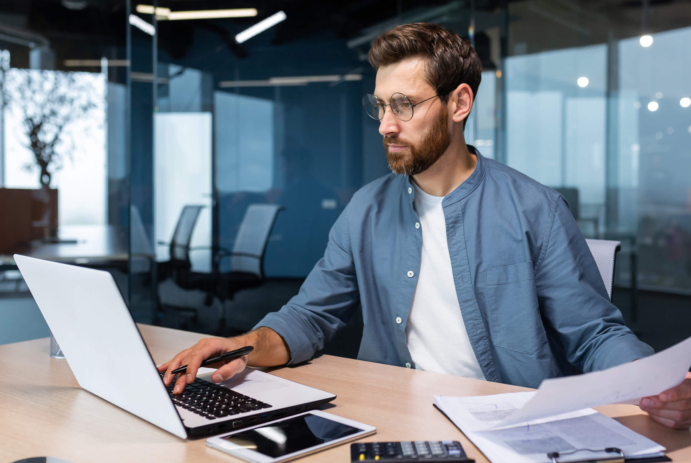 A man with glasses and a beard, wearing a blue shirt, works on a laptop at a desk with papers, a calculator, and a tablet in a modern office.