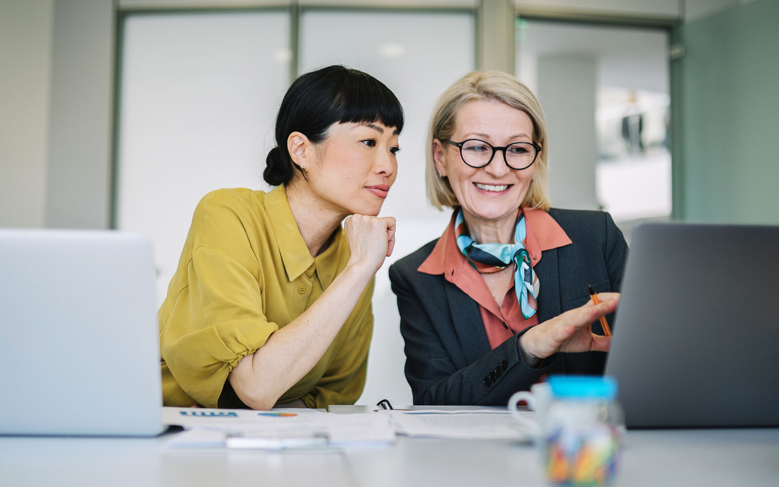 Two women collaborating over a laptop in a modern office, one in a yellow blouse and the other in a suit with a scarf, with papers and a jar of pens on the desk.